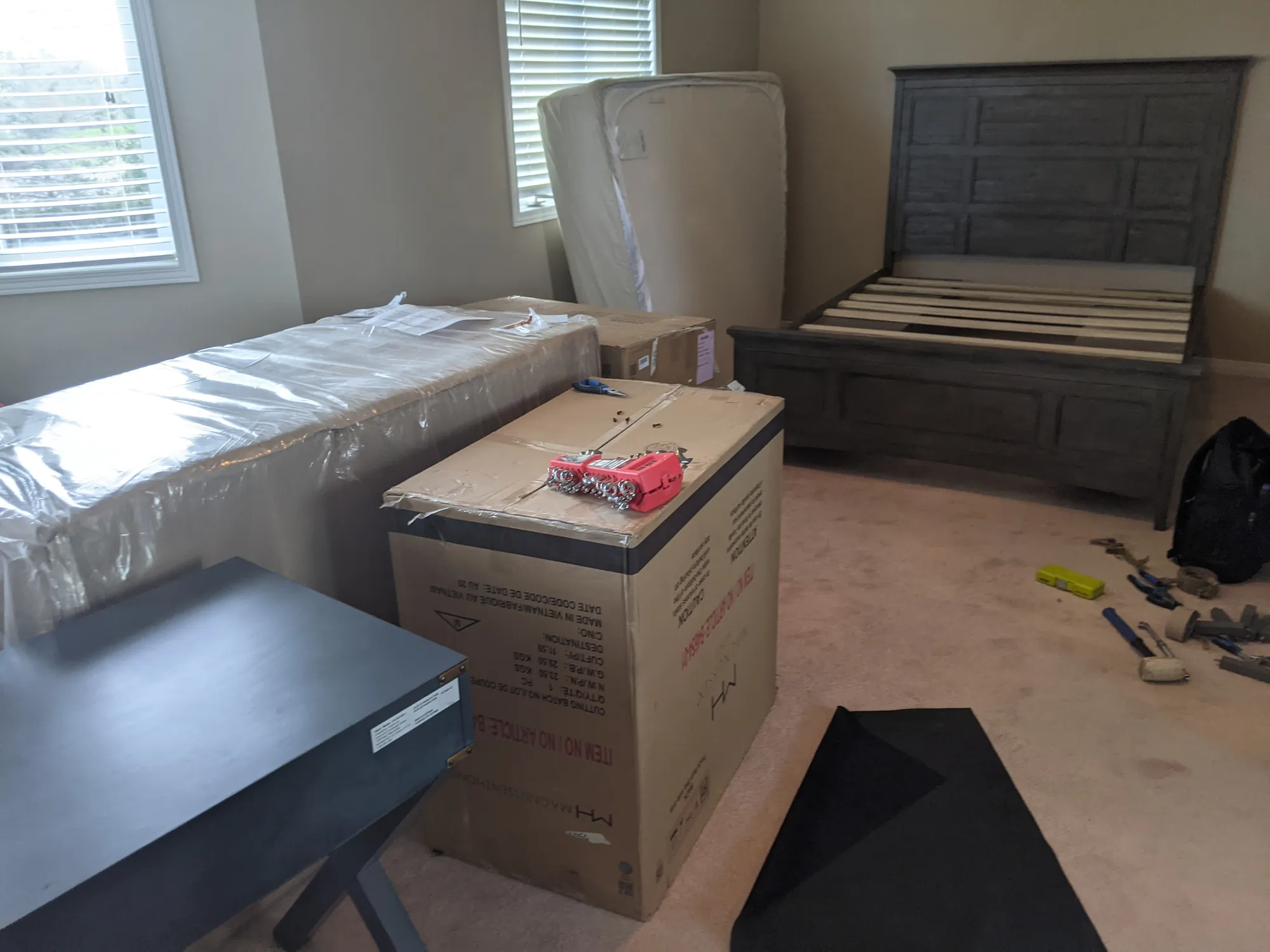 A bedroom part-way through a move: a bed frame being reassembled, a wrapped mattress standing alongside and tools laid out on the floor