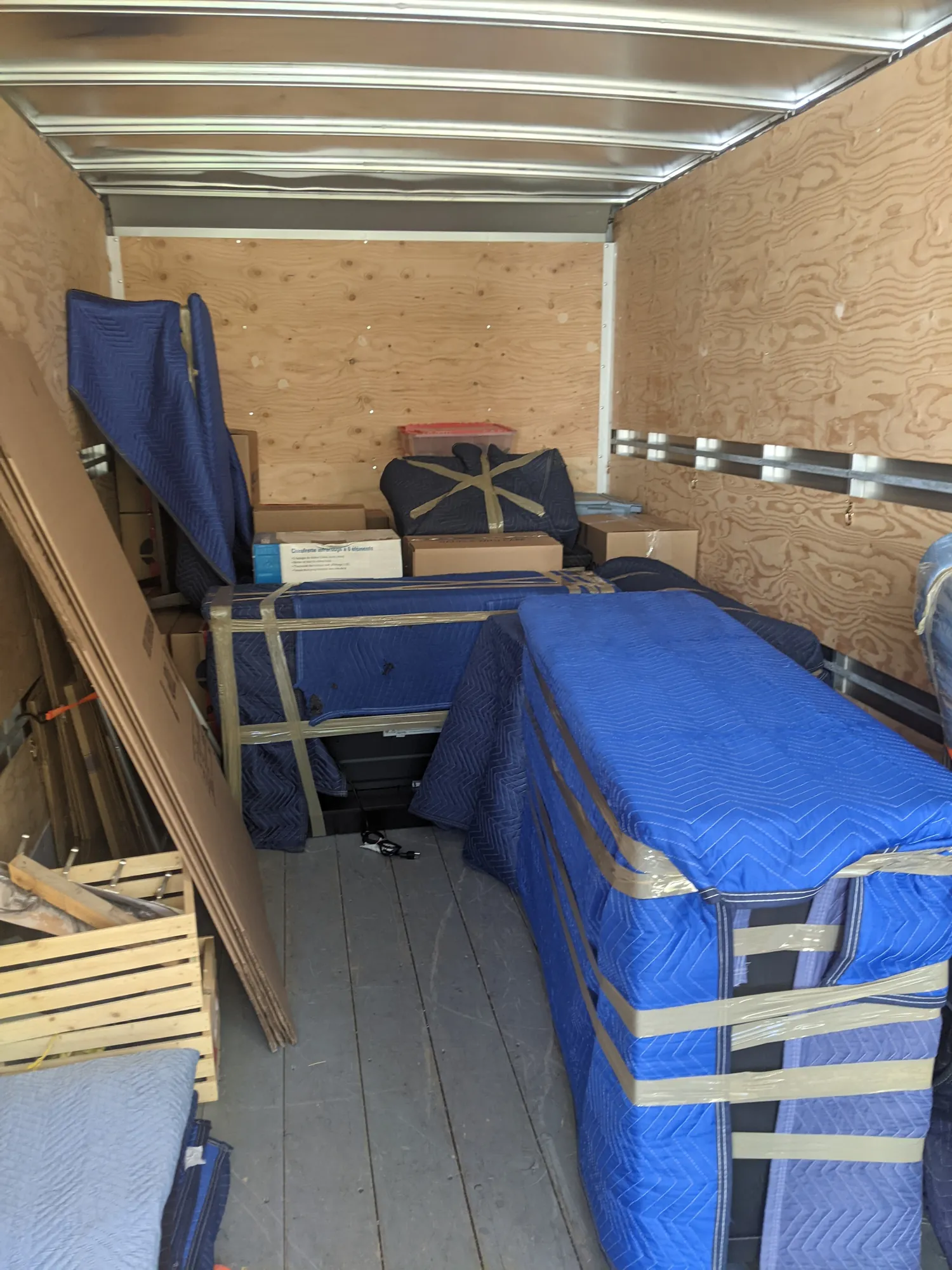 Looking into a truck box part-way through loading: furniture wrapped in blue moving blankets and taped, cartons behind, and flat cardboard and a wooden crate along one wall