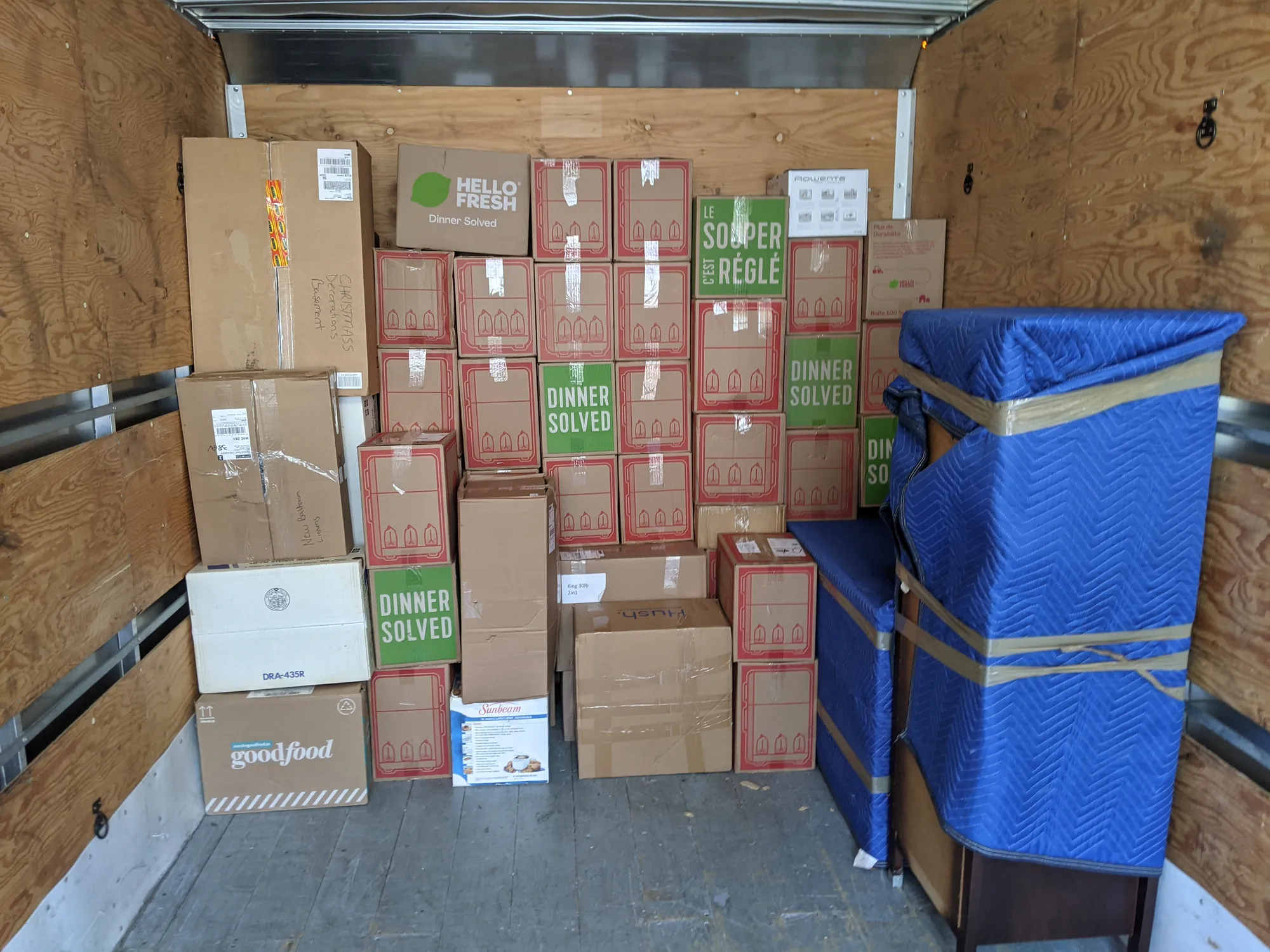 A truck box with a full wall of taped cartons stacked floor to ceiling and two pieces of furniture wrapped in blue moving blankets and taped beside them