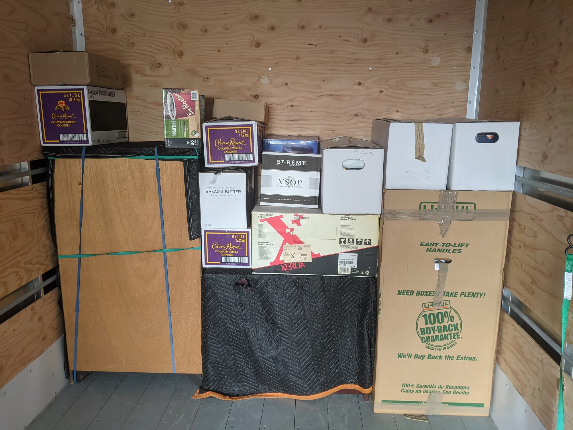 Cartons and a blanket-covered cabinet stacked against the front wall of a truck box, with a tall wardrobe carton at one side and straps holding a panel in place
