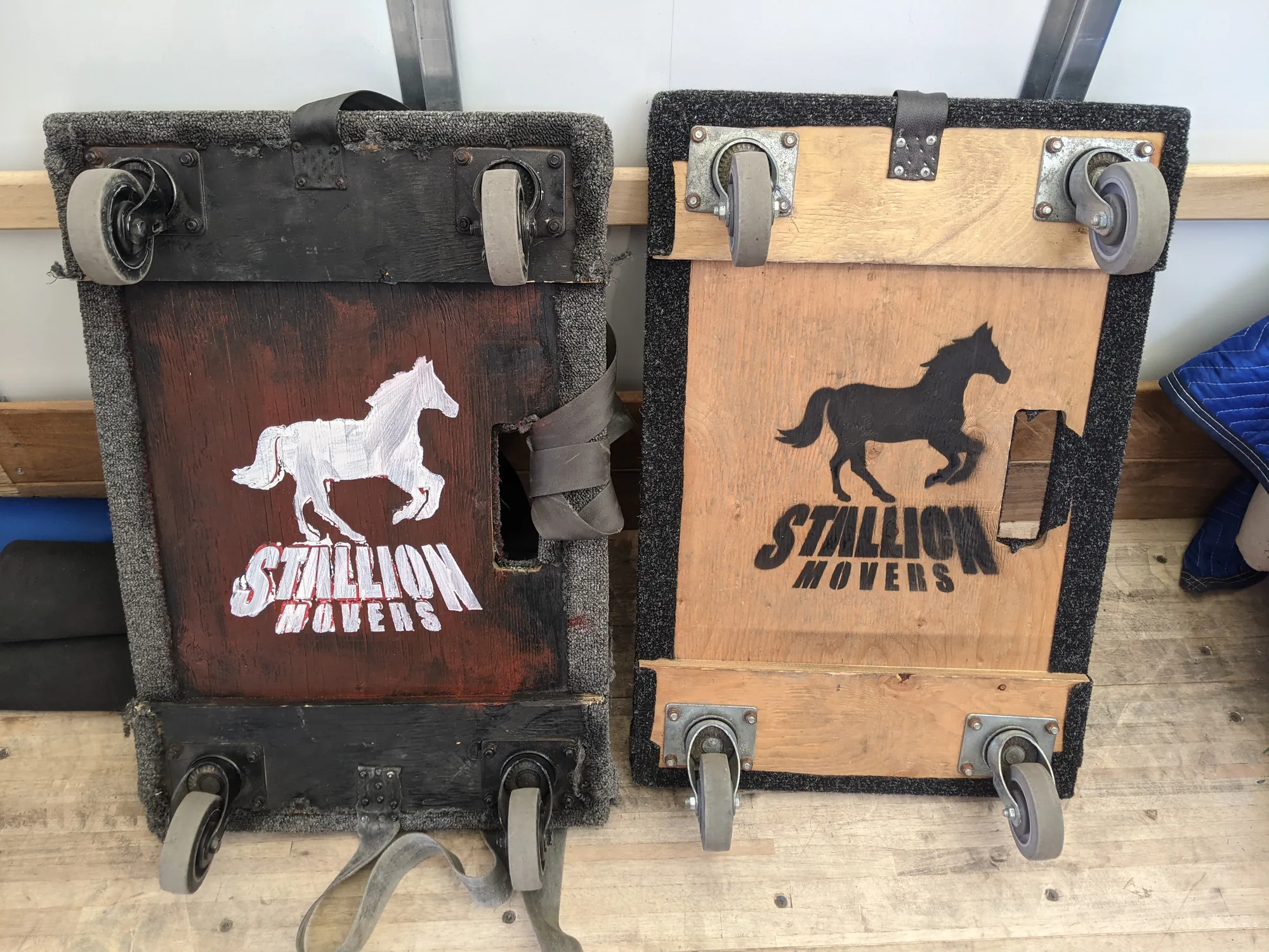 Two hand-painted Stallion four-wheel furniture dollies standing against a wall, one worn dark red and one bare wood