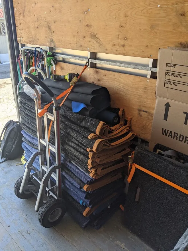A hand truck standing against a tall stack of folded moving blankets inside a truck box, with rolled floor runners, straps hung on the wall and a wardrobe carton alongside
