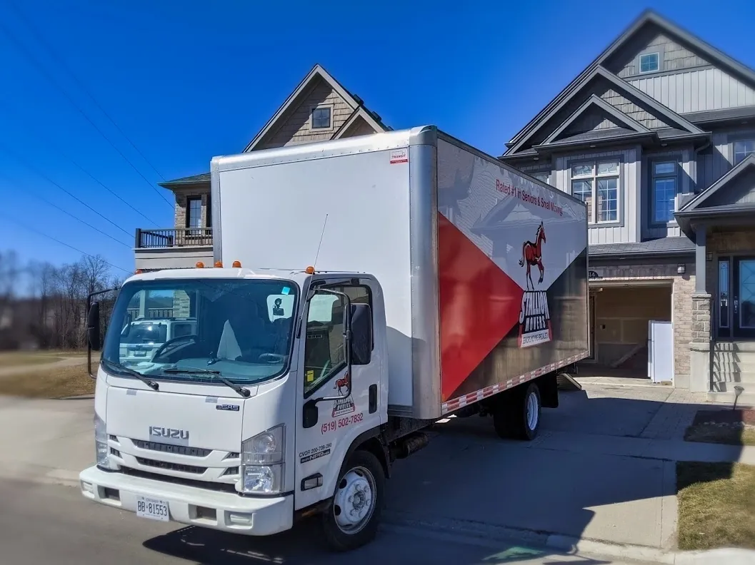 A branded Stallion box truck parked at the kerb in front of two tall new houses under a clear blue sky, with a garage door open behind it